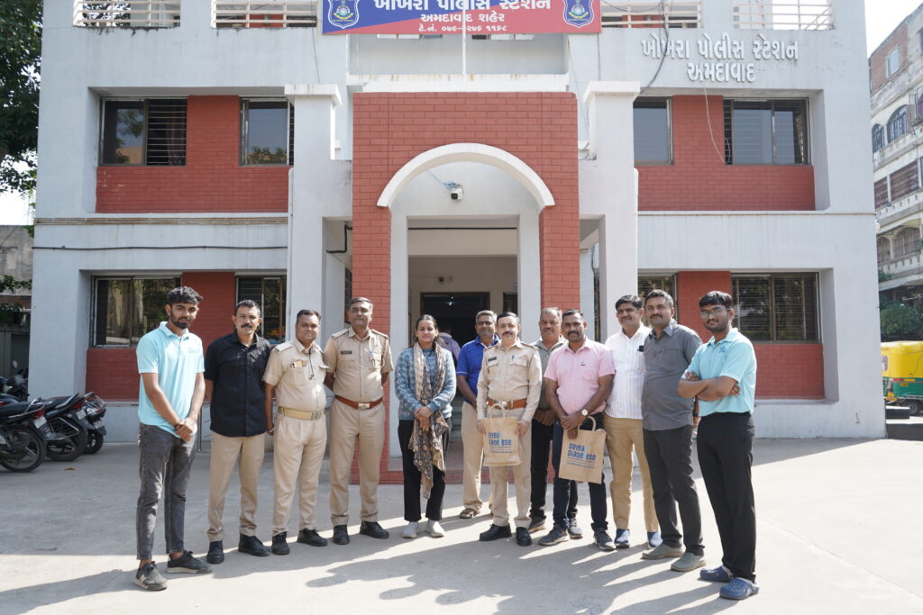 A group of approximately 11 people, including police officers in uniform and civilians, posing for a photo in front of the Amraiwadi Police Station in Ahmedabad.
