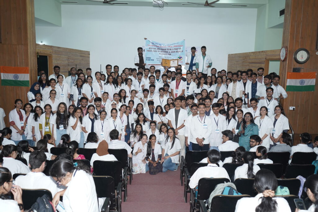 A very large group of students, mostly wearing white coats, posing for a group photo in an auditorium with Indian flags on either side of the stage.